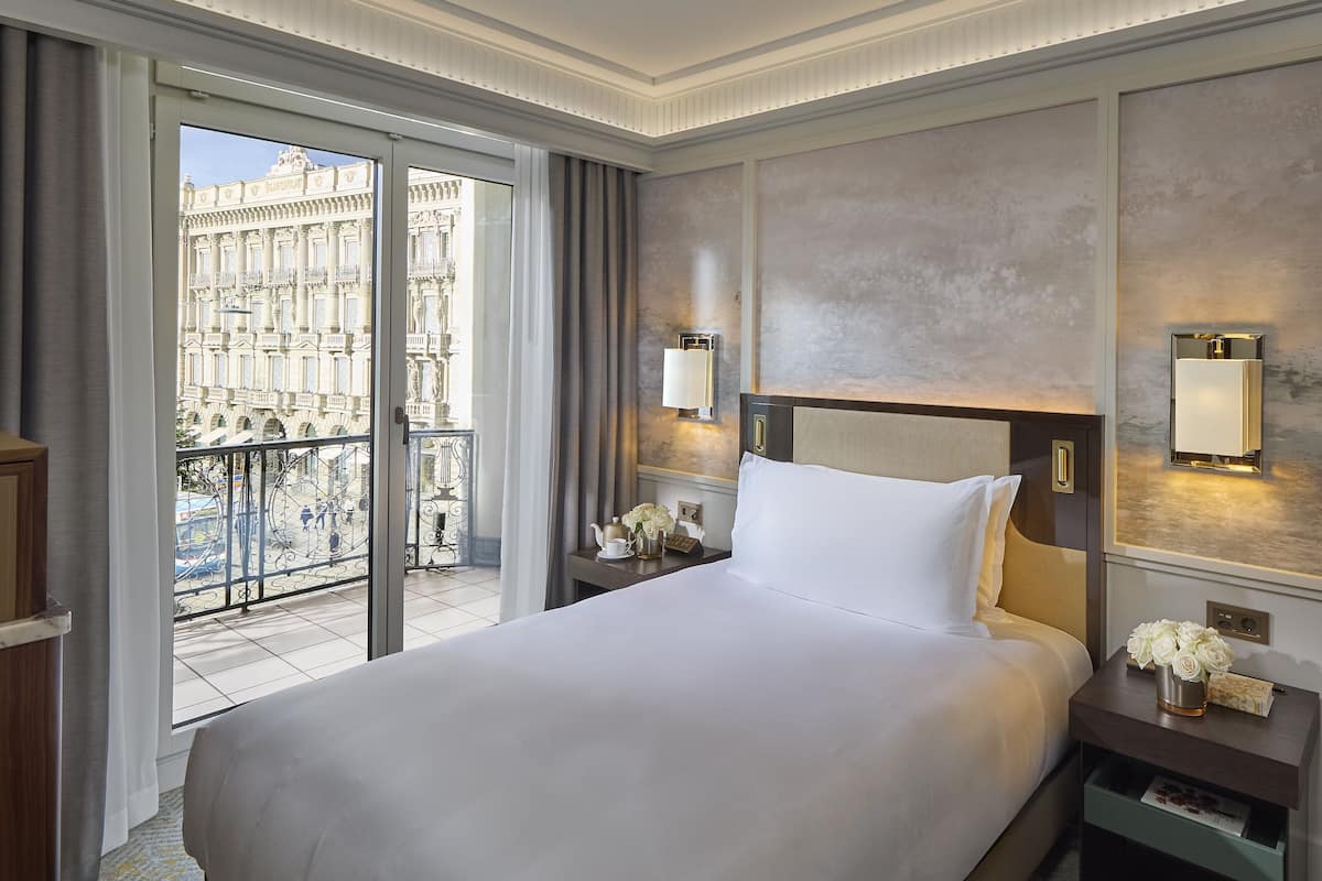 A hotel room with a large bed, a nightstand with a lamp, a view of a building through a glass door, and a balcony with a railing.