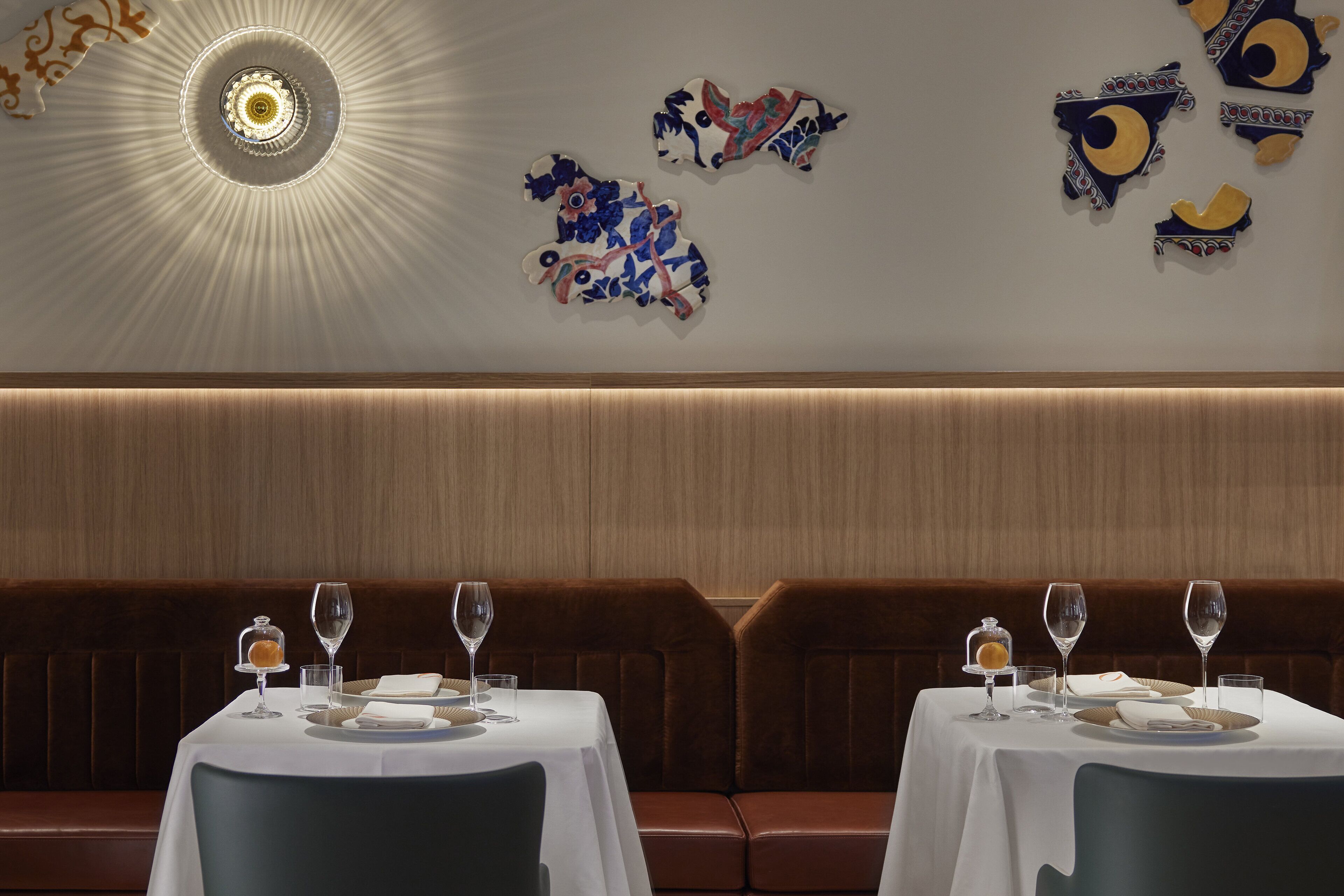 Un restaurant avec deux tables dressées pour le dîner, chacune recouverte d’une nappe blanche et agrémentée de verres à vin. Les murs sont décorés d’œuvres d’art et un luminaire est suspendu au plafond.