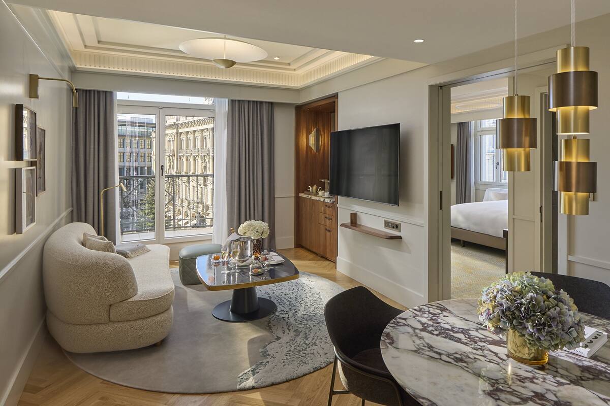 A modern hotel room with a living area, a dining table, a sofa, a TV, and a balcony with a city view.