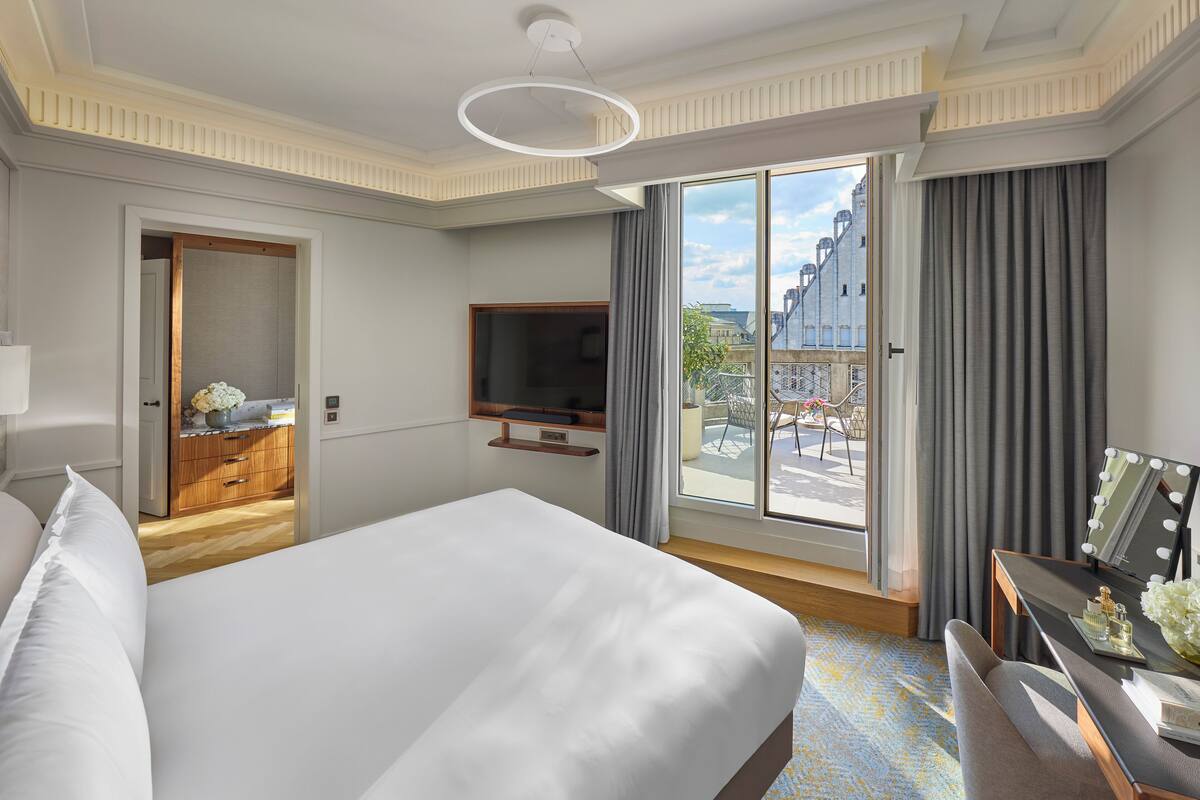 A hotel room with a large bed, a TV mounted on the wall, a balcony with a view of the city, and a desk with a mirror and toiletries.
