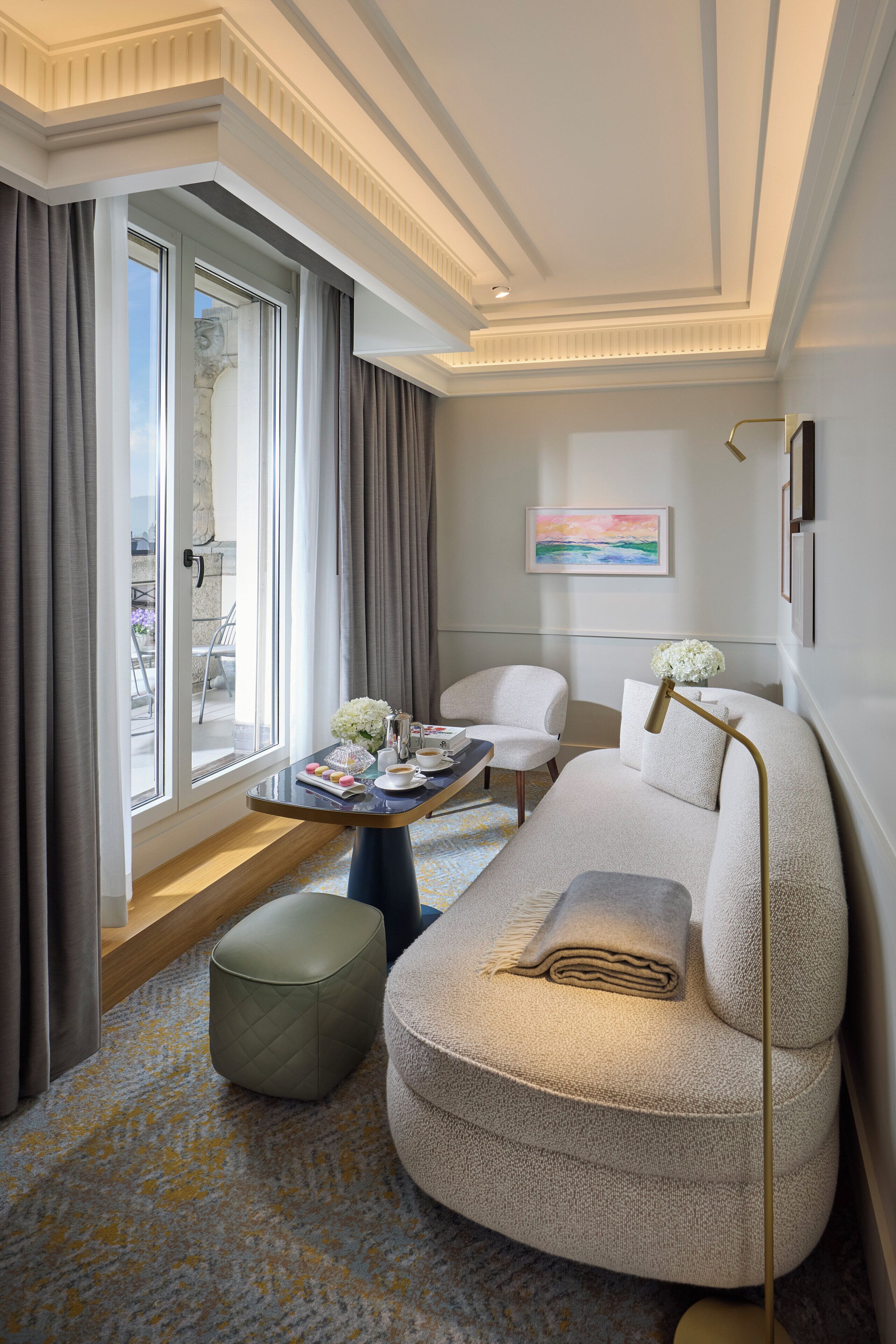 A modern hotel room with a large window, a sofa, a coffee table, and a painting on the wall.