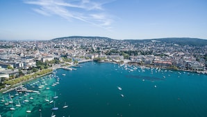 An aerial view of a coastal city with a marina filled with boats, surrounded by dense urban buildings and hills in the background. - Mandarin Oriental Savoy, Zurich (Zürich)