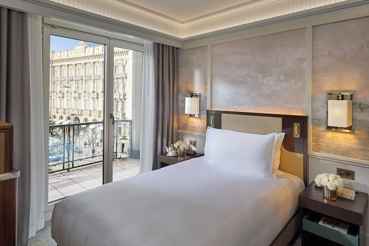 A hotel room with a large bed, a nightstand with a lamp, a view of a building through a glass door, and a balcony with a railing.
