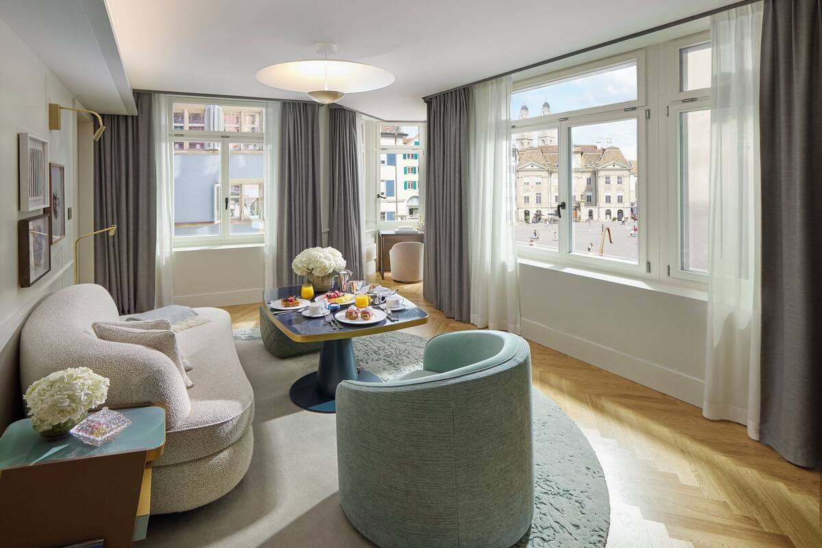 A modern hotel room with a large window, a dining area with breakfast, a sofa, and a round chair.
