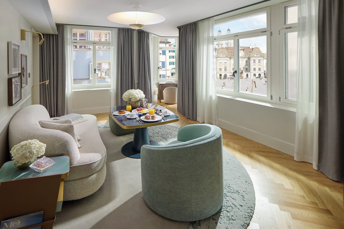 A modern hotel room with a large window, a dining area with breakfast, a sofa, and a round chair.