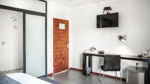 A modern hotel room with a shower, a wooden door, a desk with a chair, a television, and a minibar. - Yalla Yalla Boutique Hotel (Emalahleni)