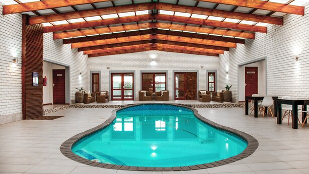 An indoor swimming pool with a wooden ceiling and white brick walls. There are seating areas with chairs and tables around the pool. - Yalla Yalla Boutique Hotel (Emalahleni)