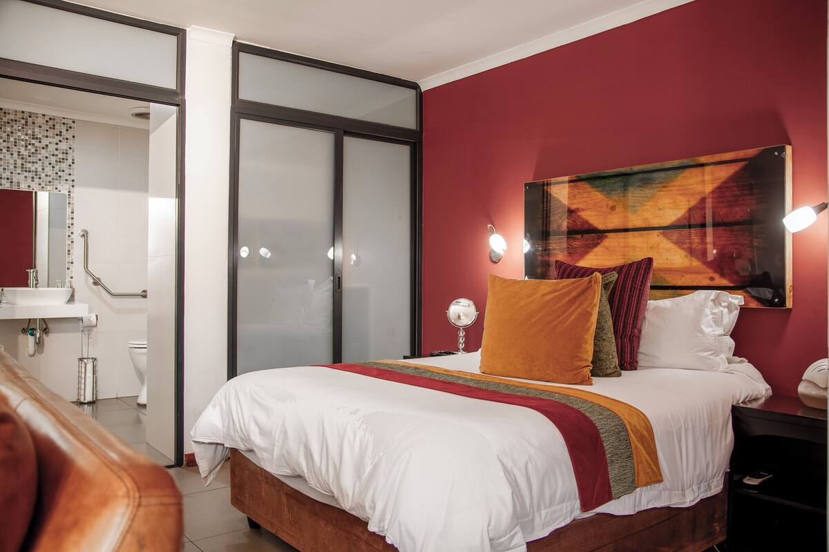 Habitación de hotel con cama, mesita de noche, lámpara, espejo, ventana y baño con lavamanos e inodoro.
