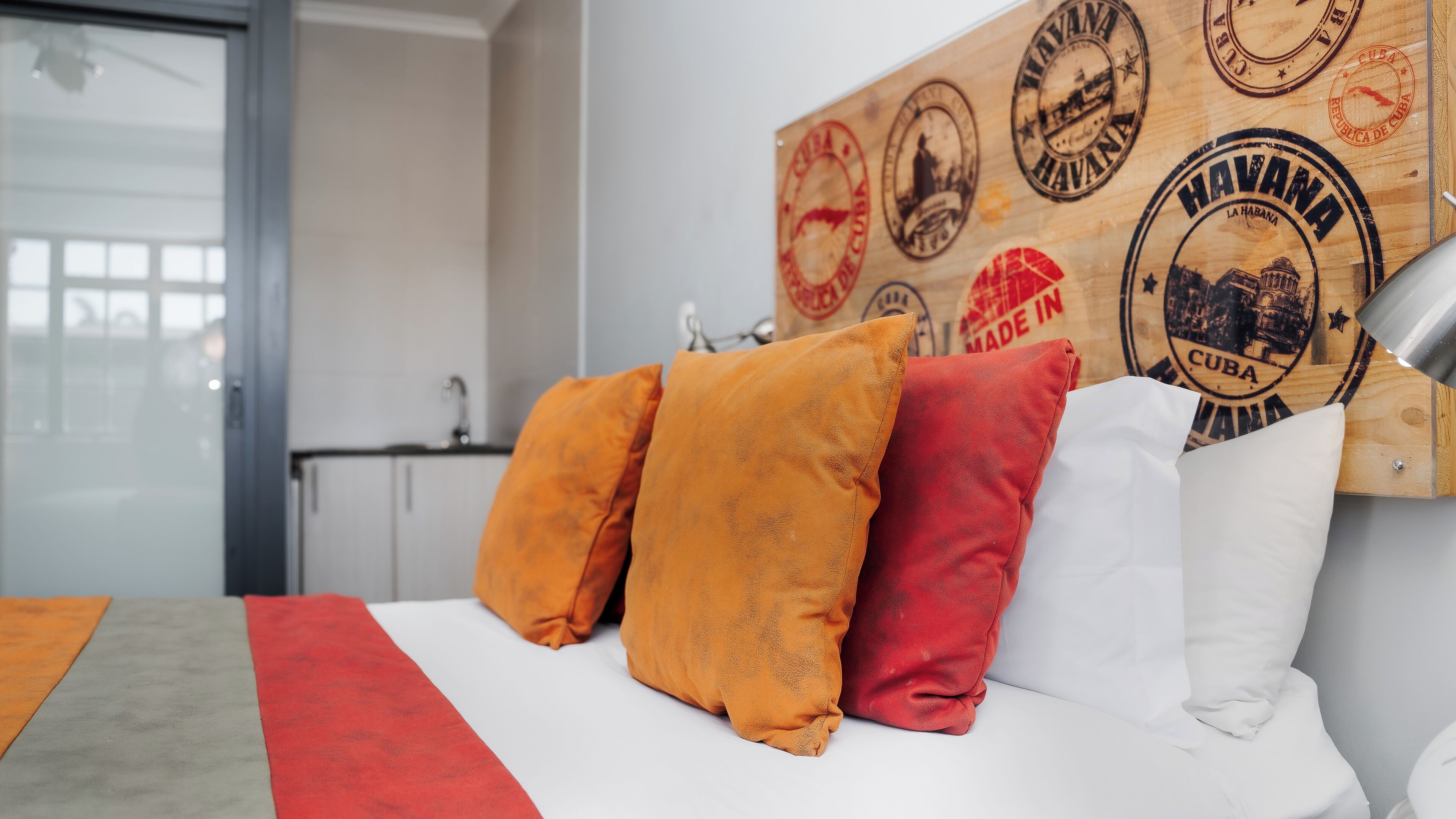 Ein Hotelzimmer mit Bett, Kissen, einer Wand voller Briefmarken, einem Fenster und einem Waschbecken.