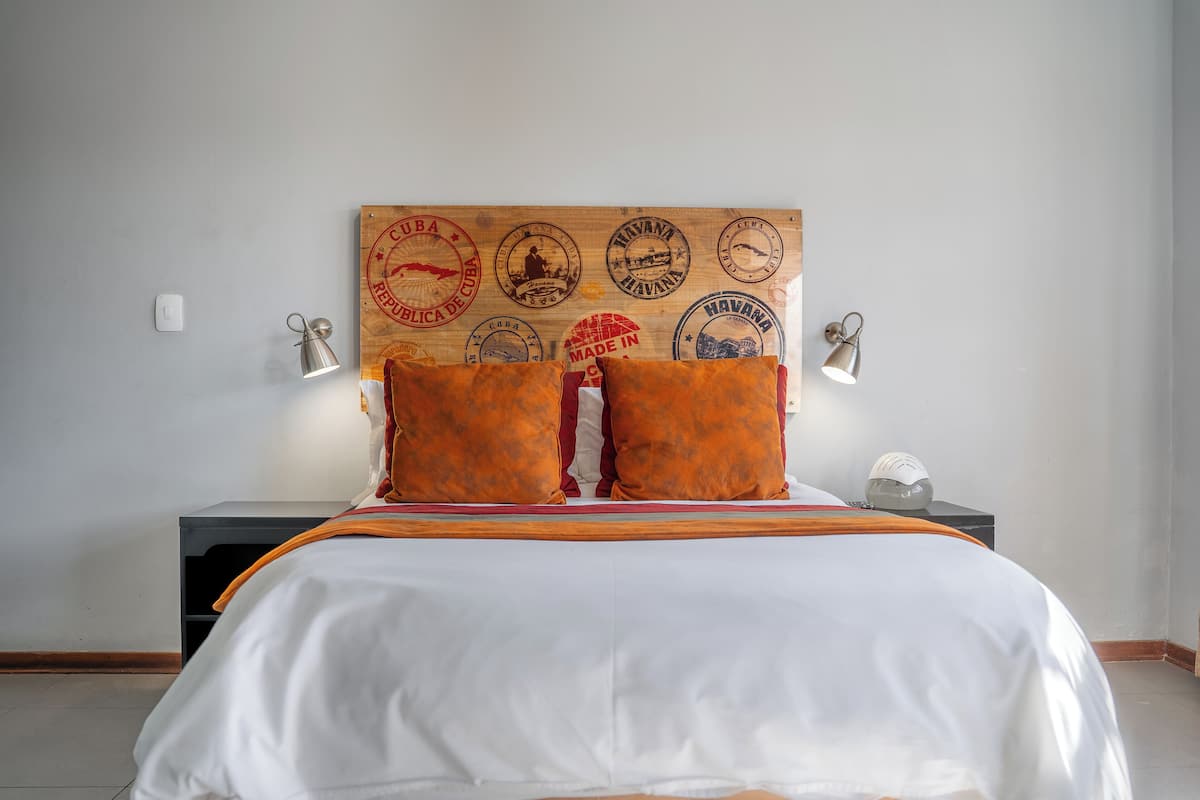 Habitación de hotel con cama, dos almohadas, cabecera adornada con sellos antiguos, lámparas de noche y mesita.