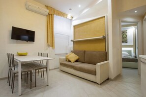 A compact living space with a sofa, a dining table with chairs, a flat-screen TV mounted on the wall, and a bed in the adjacent room. - Gold Vatican Apartment (Rome)