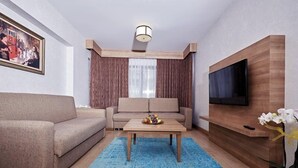A modern living room with a sofa, a coffee table, a TV mounted on a wooden wall, and a large window with curtains. - Bursa Suites Apart Hotel (Bursa)