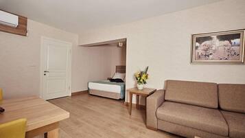 Habitación de hotel con piso de madera, sofá color beige, una pequeña mesa con un florero y una cama con manta azul, además de un cuadro en la pared.