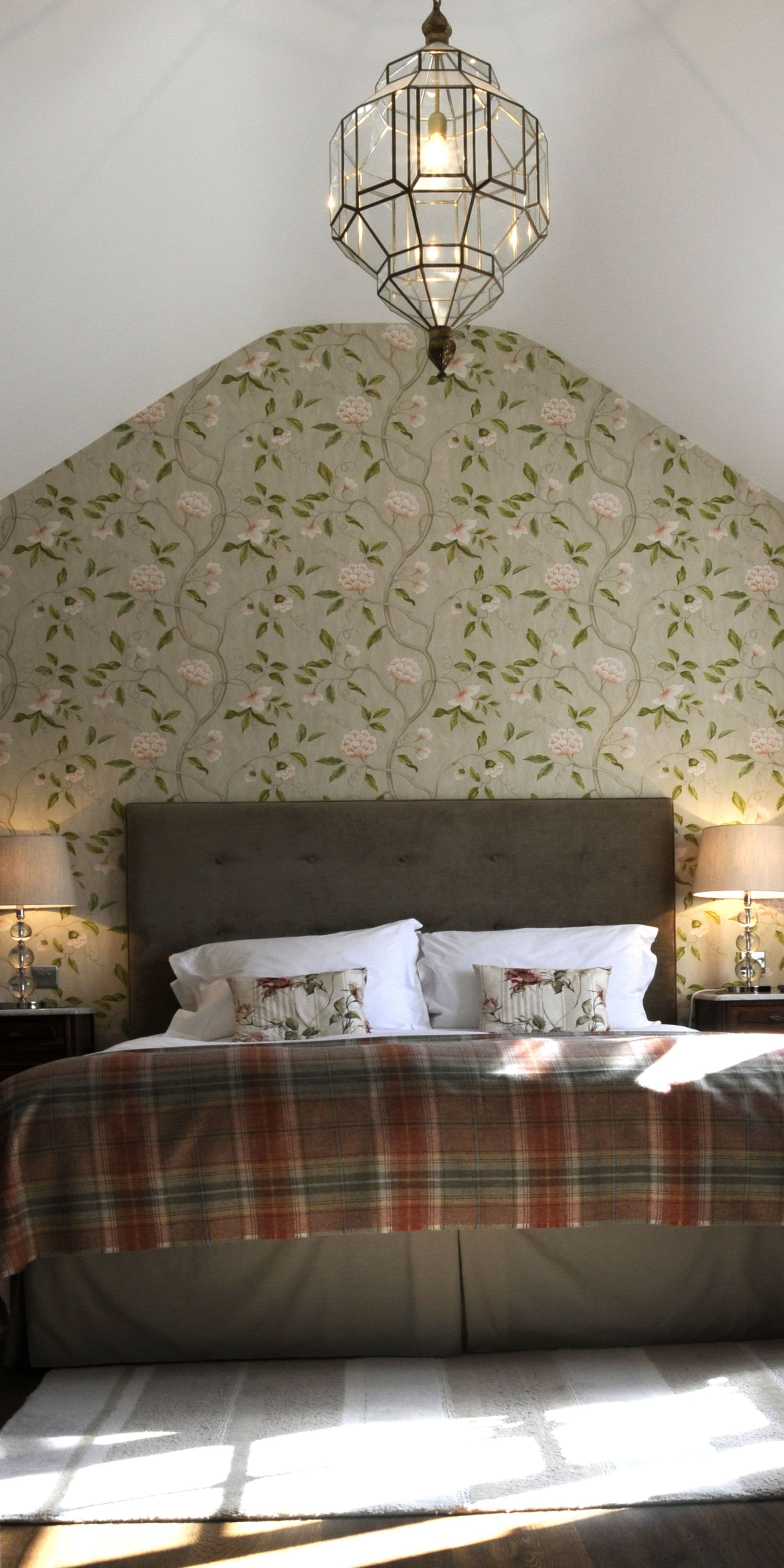 Ein Schlafzimmer mit einem großen Bett, floralem Wandpapier, einem hölzernen Kopfteil und einer Pendelleuchte.