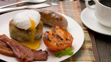 A plate with a sausage, a poached egg, bacon, and a grilled tomato on a table with a cup of coffee and salt and pepper shakers in the background.