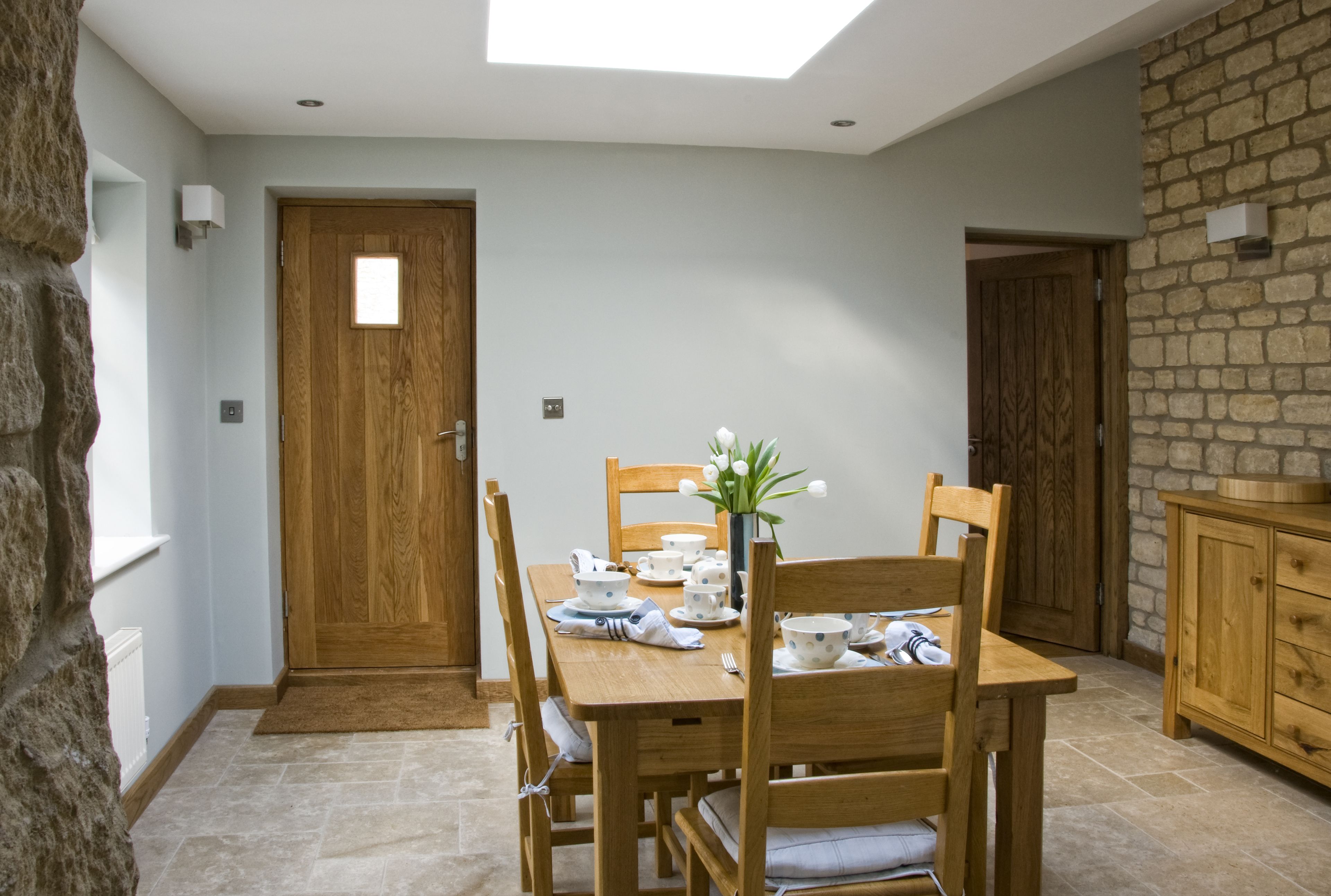 Comedor con una mesa de madera dispuesta para cuatro personas, rodeada de sillas. Un jarrón con flores está sobre la mesa, y se observa una pared de piedra y una puerta de madera al fondo.