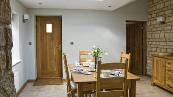 Comedor con una mesa de madera dispuesta para cuatro personas, rodeada de sillas. Un jarrón con flores está sobre la mesa, y se observa una pared de piedra y una puerta de madera al fondo.
