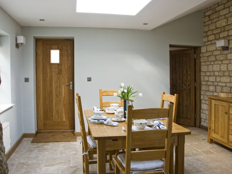 Comedor con una mesa de madera dispuesta para cuatro personas, rodeada de sillas. Un jarrón con flores está sobre la mesa, y se observa una pared de piedra y una puerta de madera al fondo.