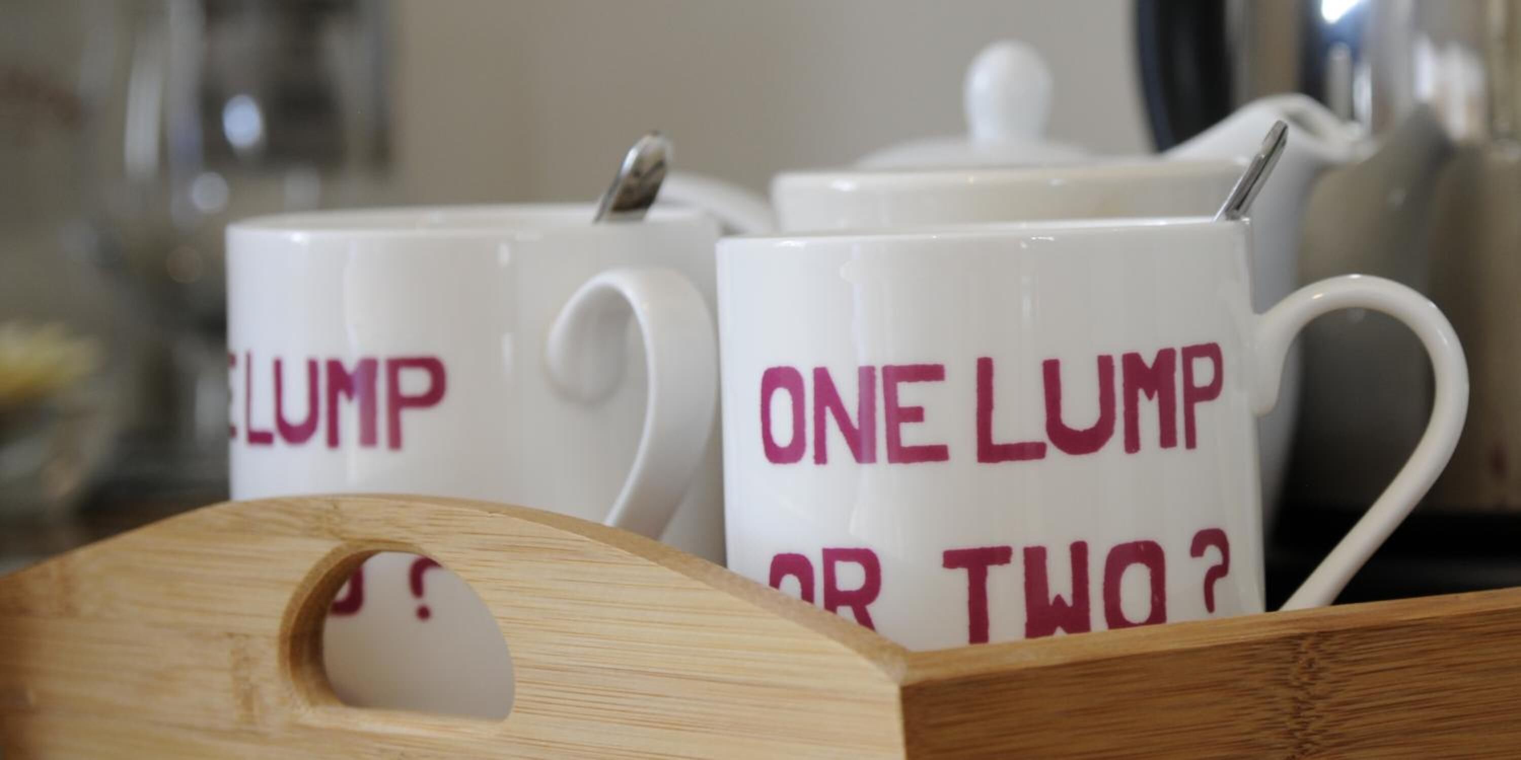 Two white mugs with "ONE LUMP OR TWO?" printed on them, placed on a wooden tray.