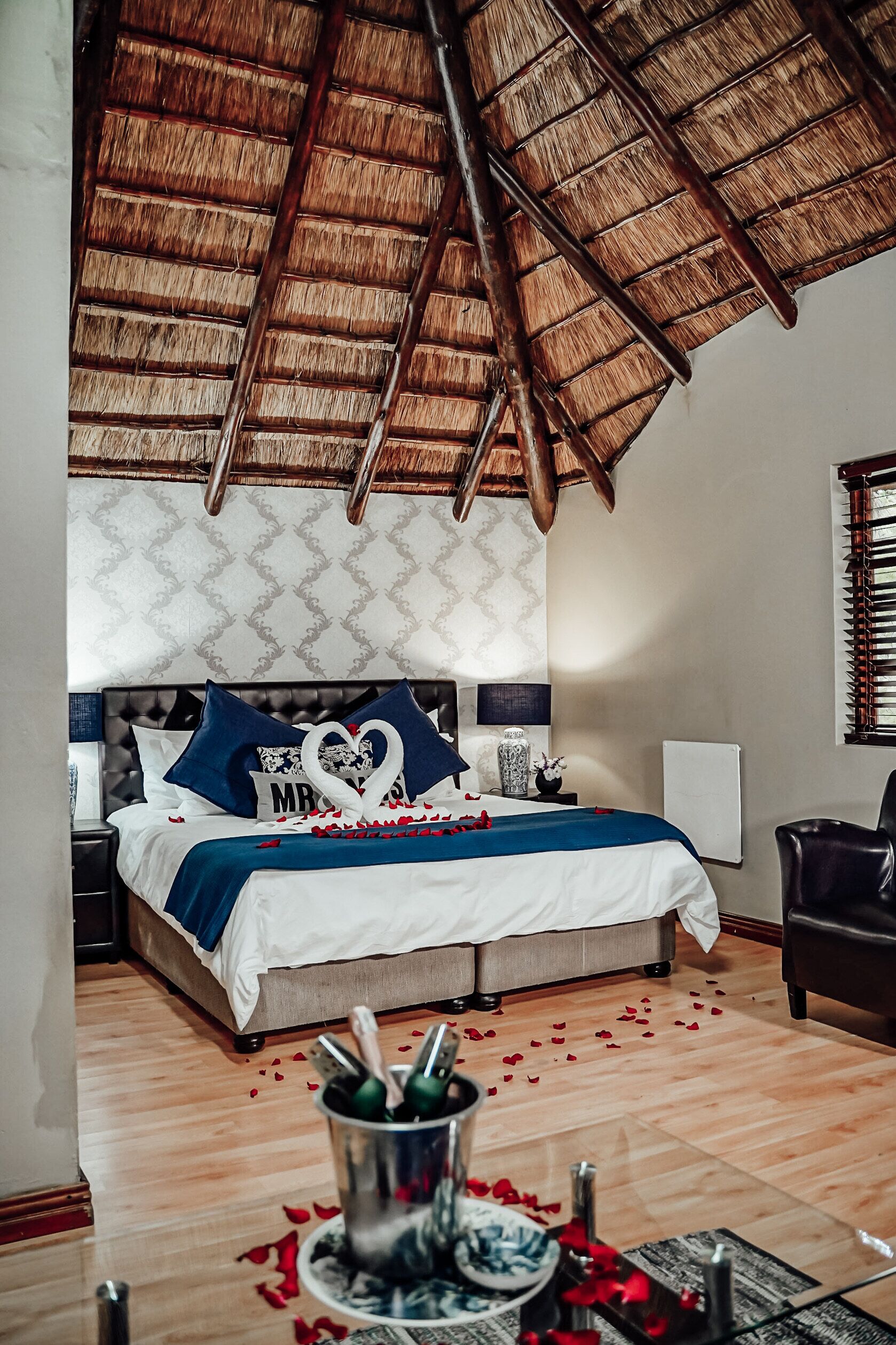 A bedroom with a bed, a nightstand, a bedside table, a window with blinds, and a ceiling with a thatched roof.