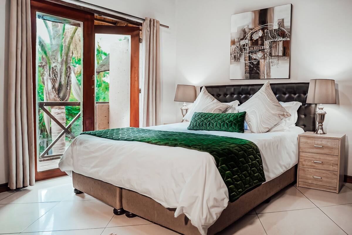 Une chambre à coucher avec un grand lit, deux tables de chevet, une fenêtre avec des rideaux et une vue sur la verdure.