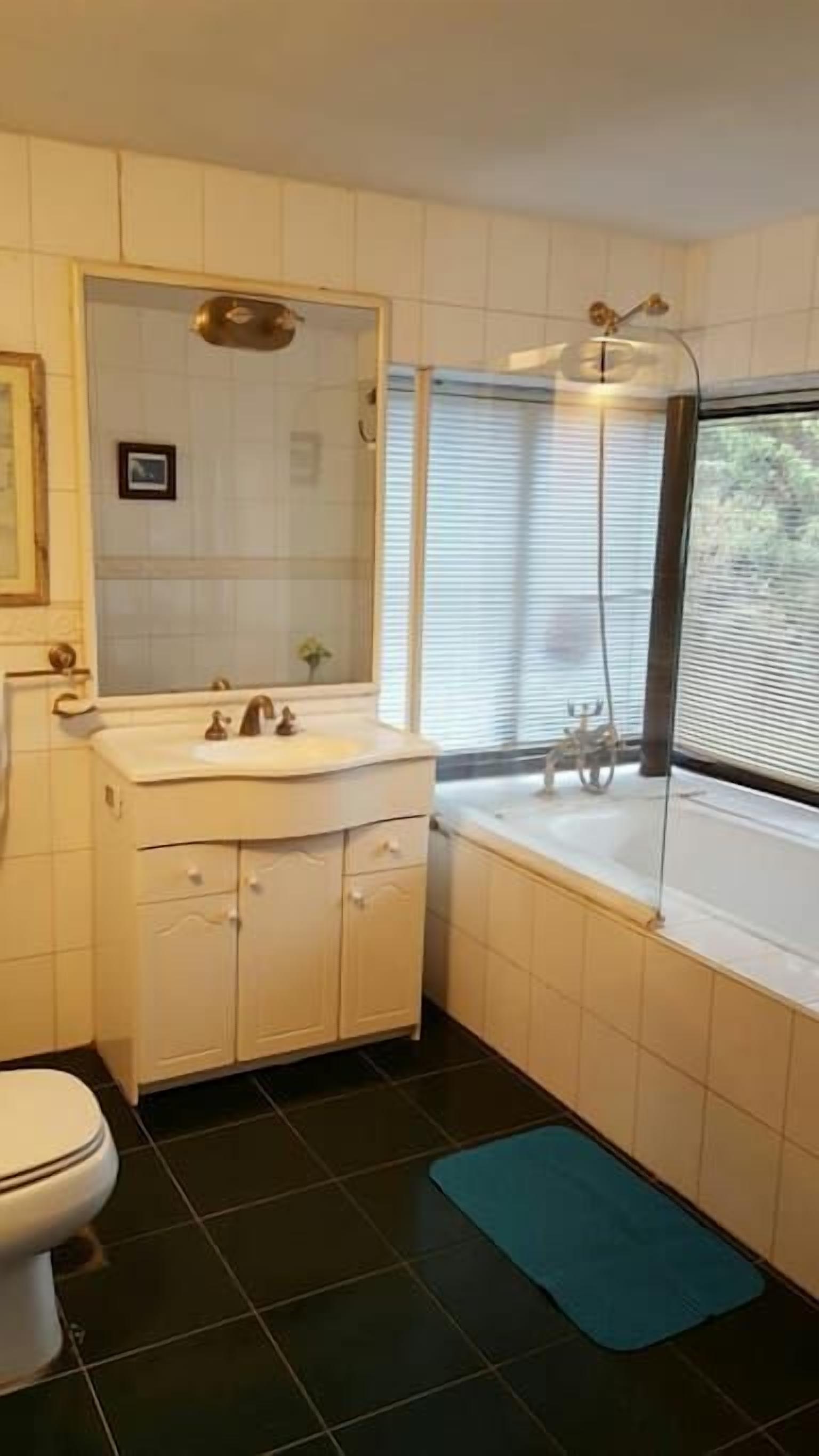 A bathroom with a white sink, mirror, and toilet. There is a window with blinds and a shower area with a glass door.