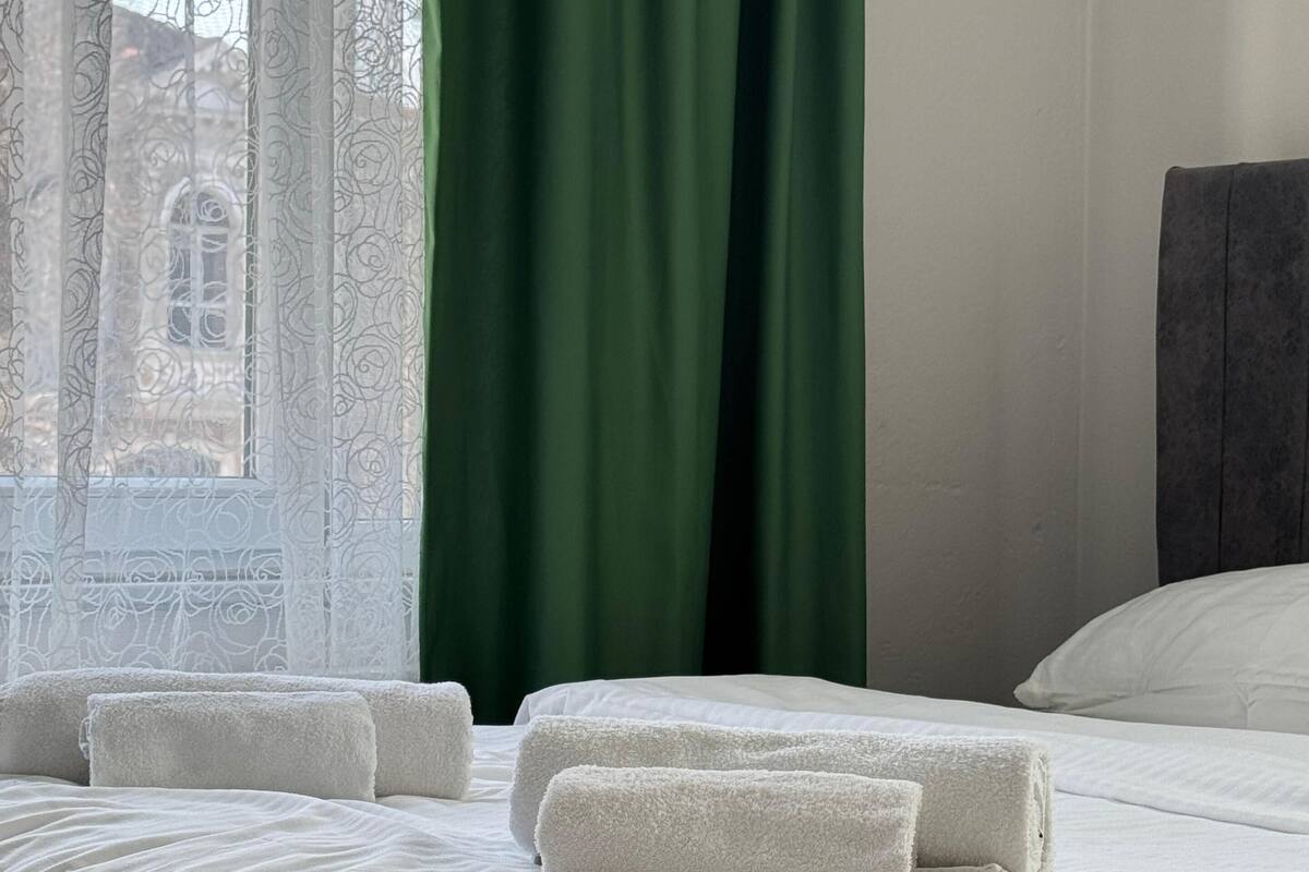 Una cama bien hecha con sábanas blancas y dos toallas enrolladas, una cortina verde y una ventana con cortinas de encaje.