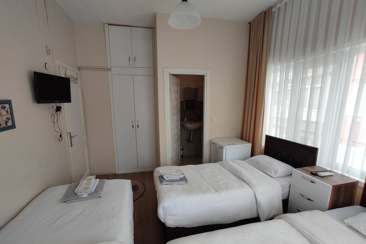 Habitación de hotel con tres camas individuales, un escritorio con silla, un refrigerador pequeño y una televisión instalada en la pared.