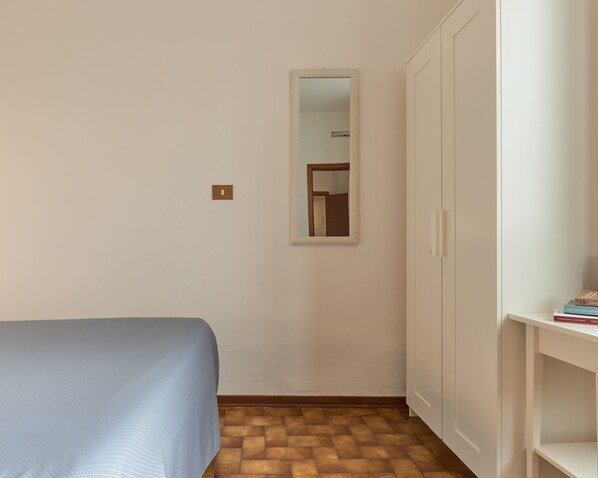 Une petite pièce simple, avec un lit, une armoire, un miroir et une étagère remplie de livres.
