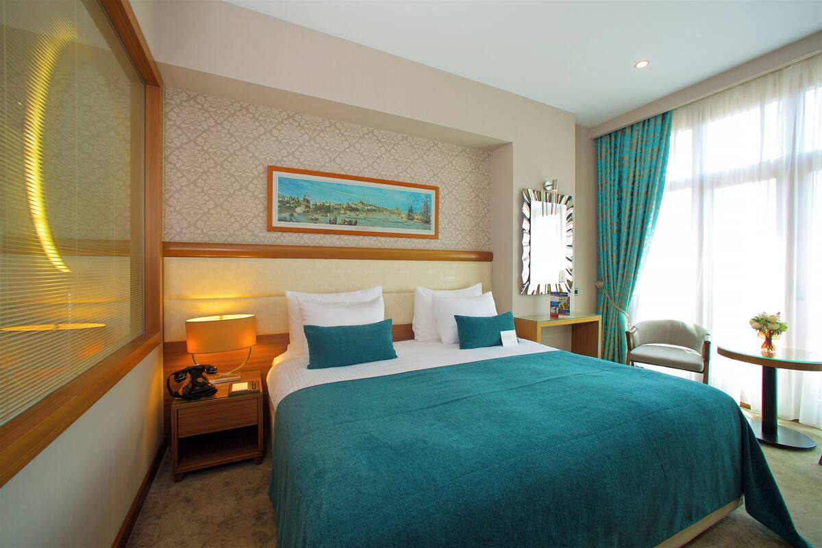 A hotel room with a large bed, a nightstand with a lamp, a chair, a small table with a plant, and a painting on the wall.