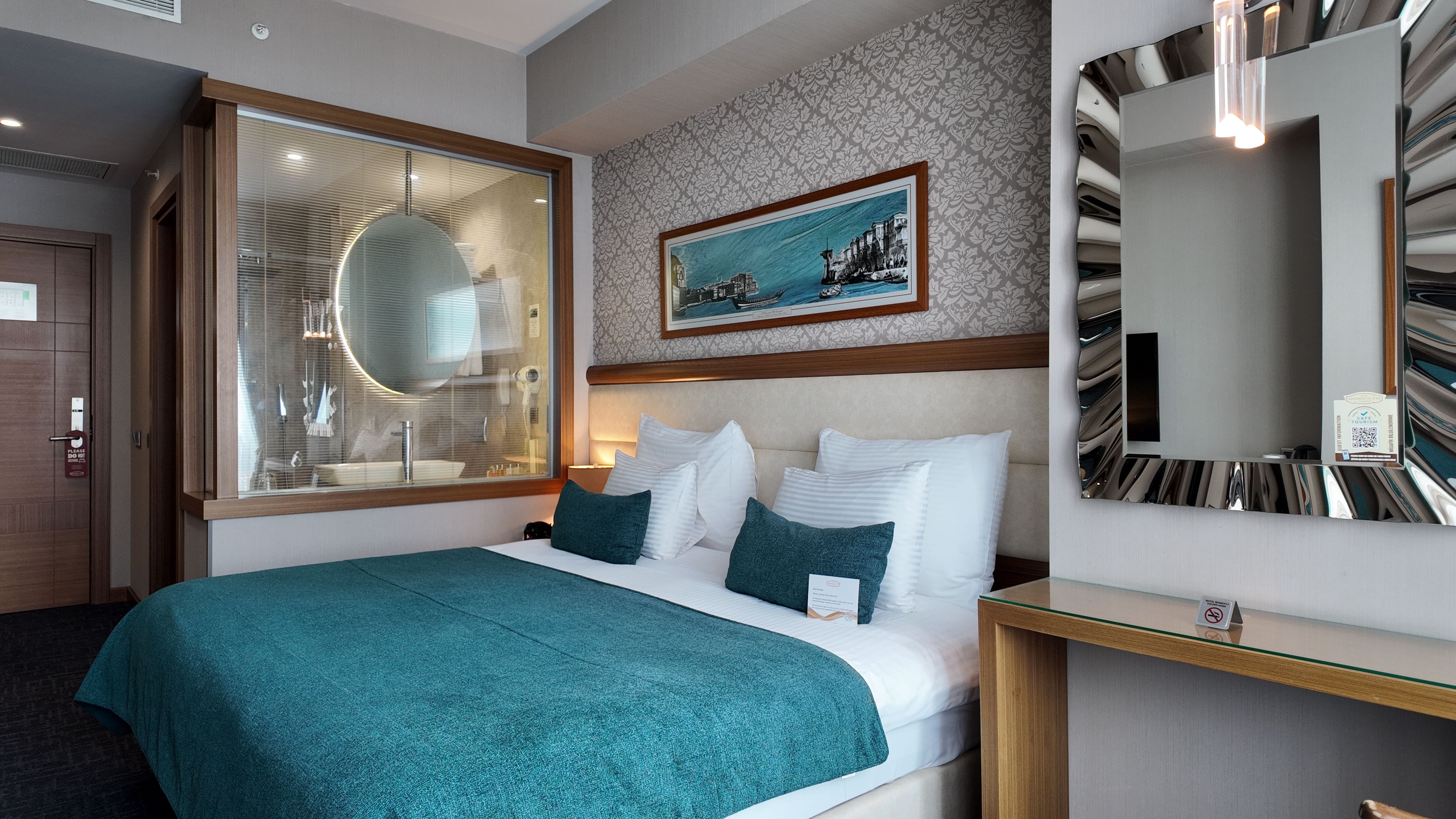 A hotel room with a large bed, a window with blinds, a painting on the wall, and a mirror with a reflection.