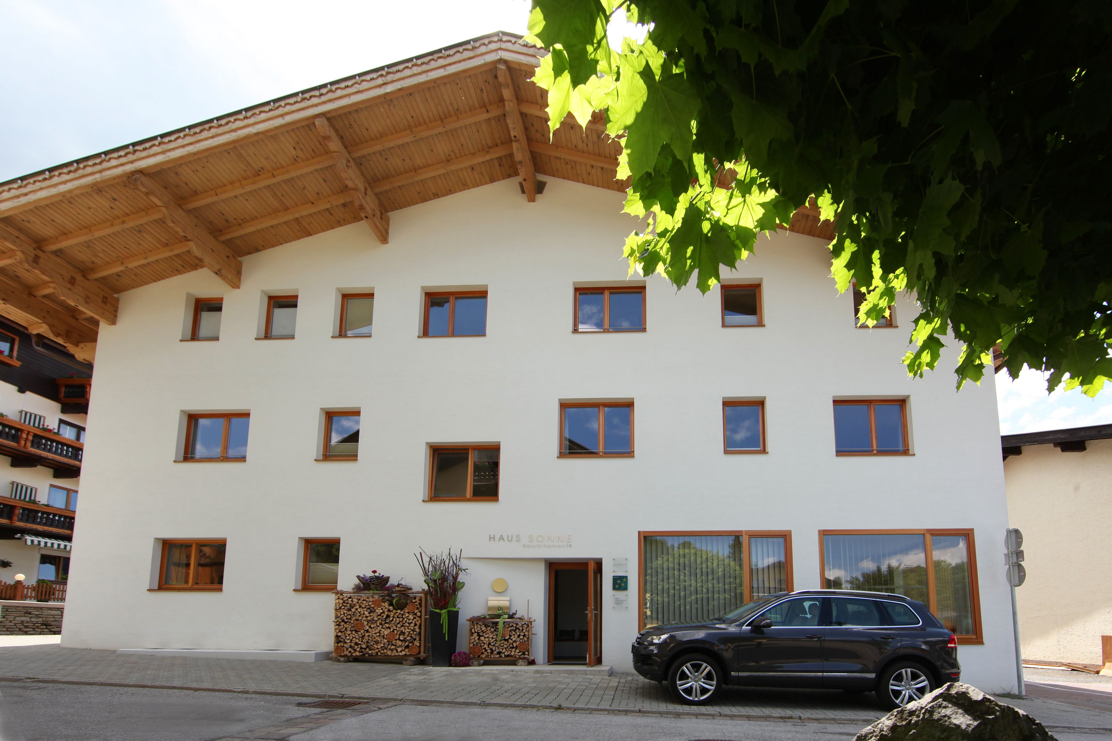 Un edificio de varios pisos con fachada blanca y vigas de madera en el techo. Se observan varias ventanas, un coche estacionado adelante y un letrero que dice “HAUS SONNE”.