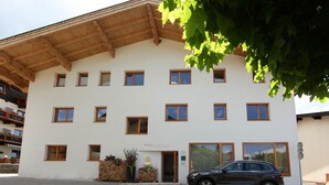 Un immeuble  à plusieurs étages, doté d’une façade blanche et de poutres en bois sur le toit. On y voit de nombreuses fenêtres, une voiture garée devant et une enseigne sur laquelle on peut lire « HAUS SONNE ».
