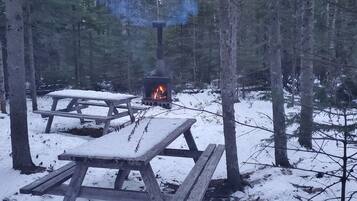 A wooden picnic table and bench set in a snowy forest with a wood-burning stove.