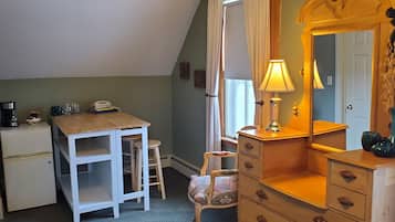 Una habitación pequeña y acogedora con un escritorio de madera, una silla, un aparador y un espejo. Hay una lámpara sobre el aparador y un refrigerador pequeño en la esquina.