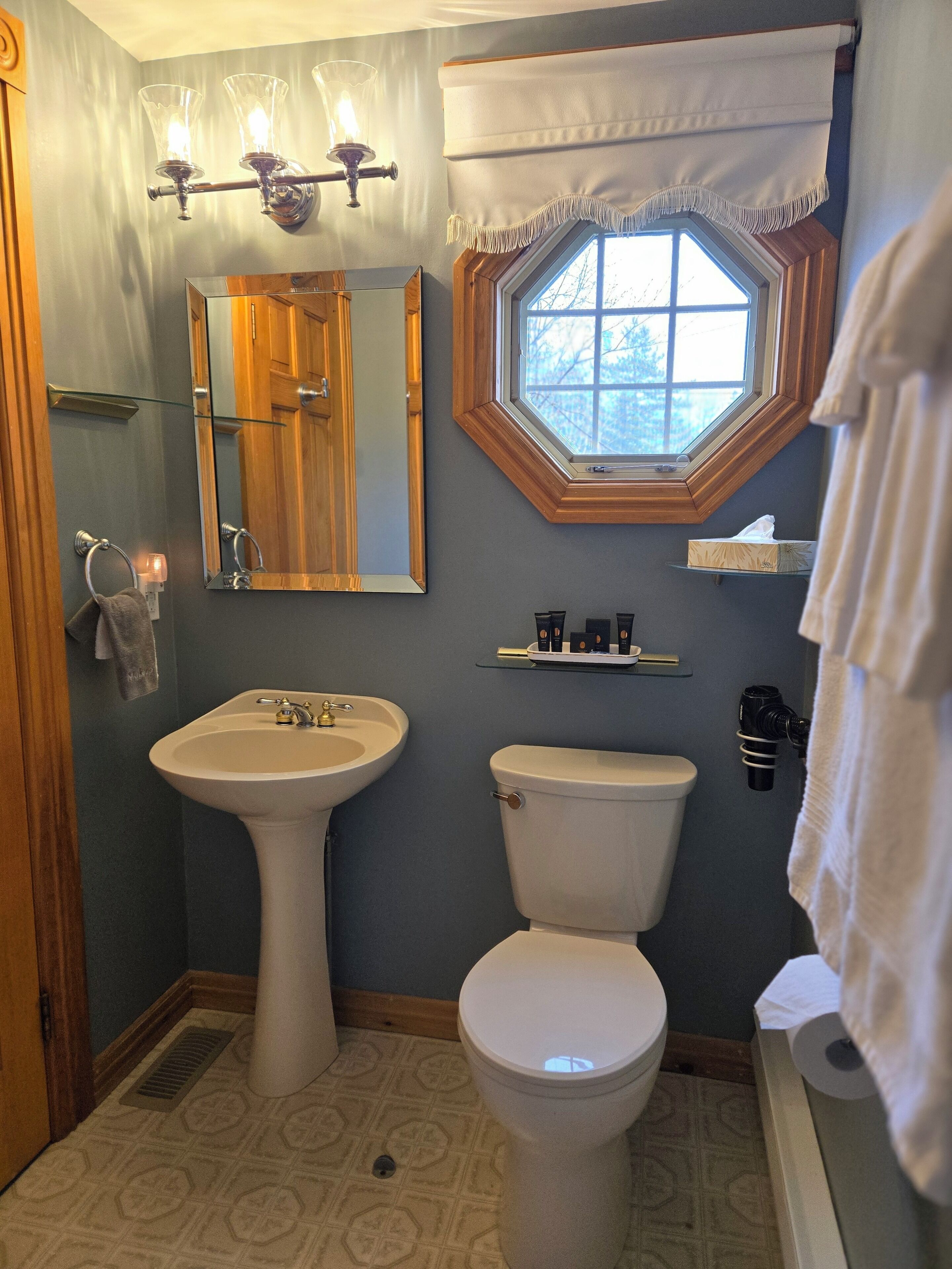 Une salle de bain avec un lavabo sur colonne, des toilettes, un miroir et une fenêtre agrémentée d’un rideau blanc.