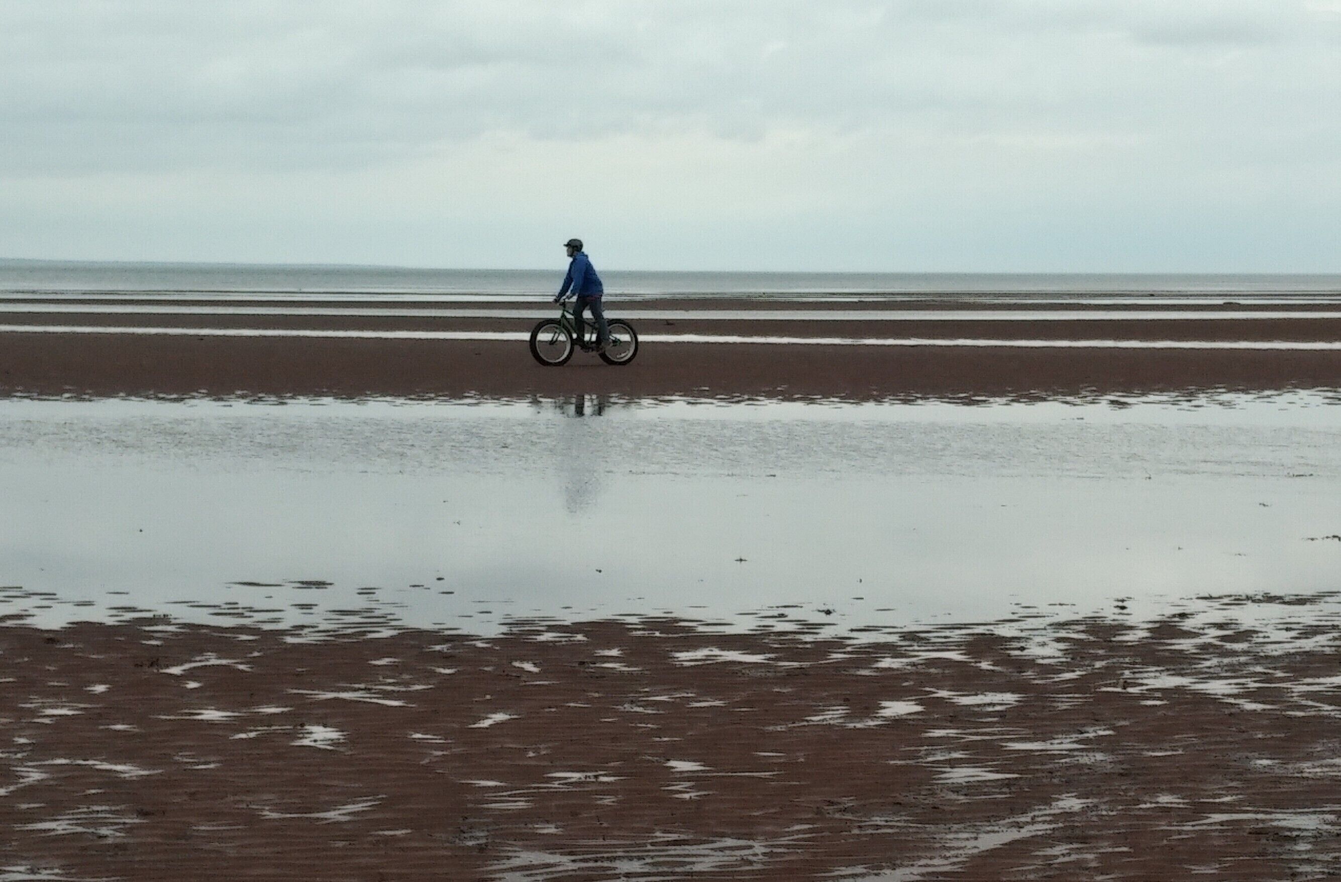 Eine Person fährt Fahrrad am feuchten Strand, im Hintergrund ein bewölkter Himmel.