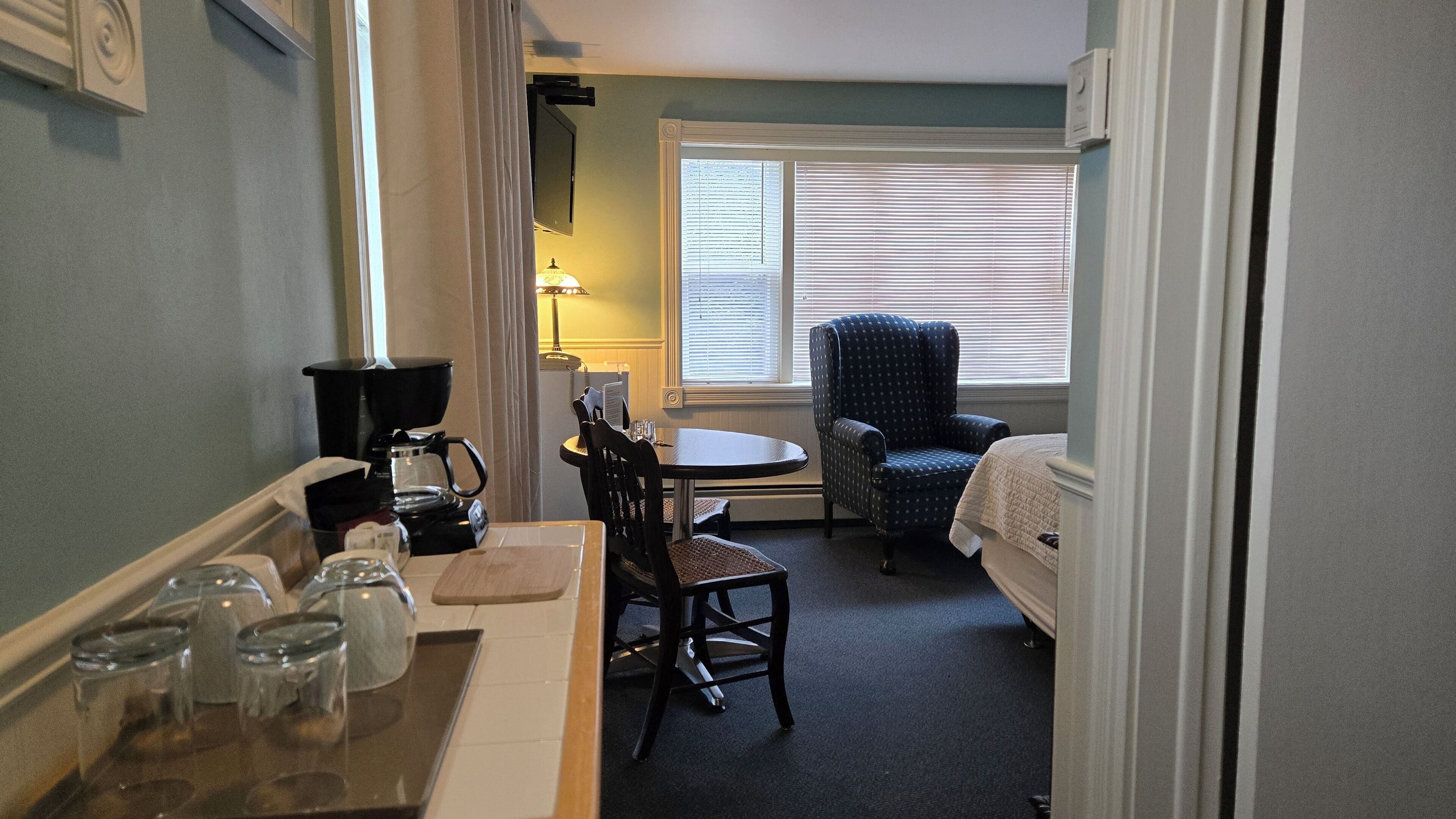 A hotel room with a dining table, chairs, a coffee maker, and a chair by the window.