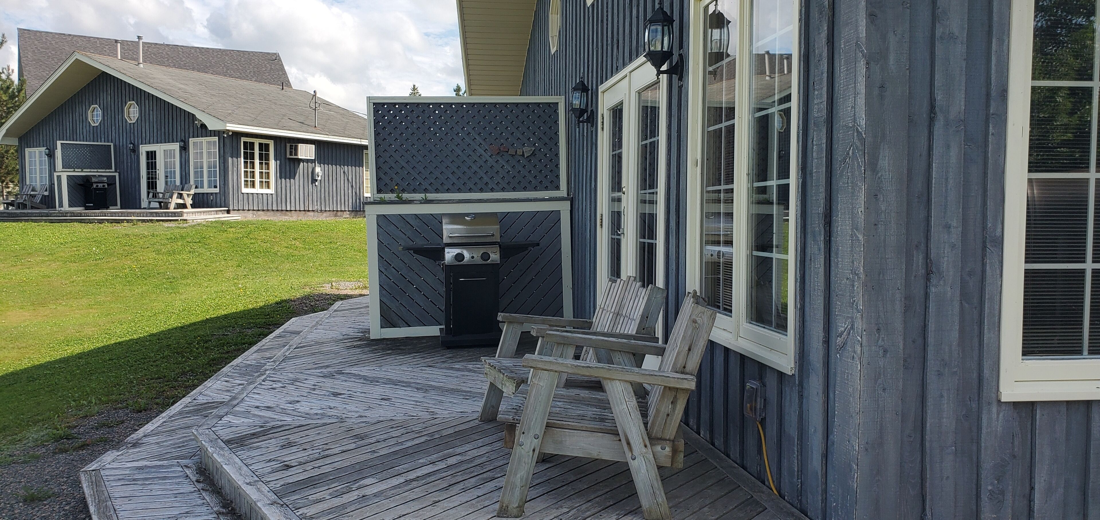 Een terras met een barbecue en stoelen, een huis met een blauwe gevel, en een gazon.