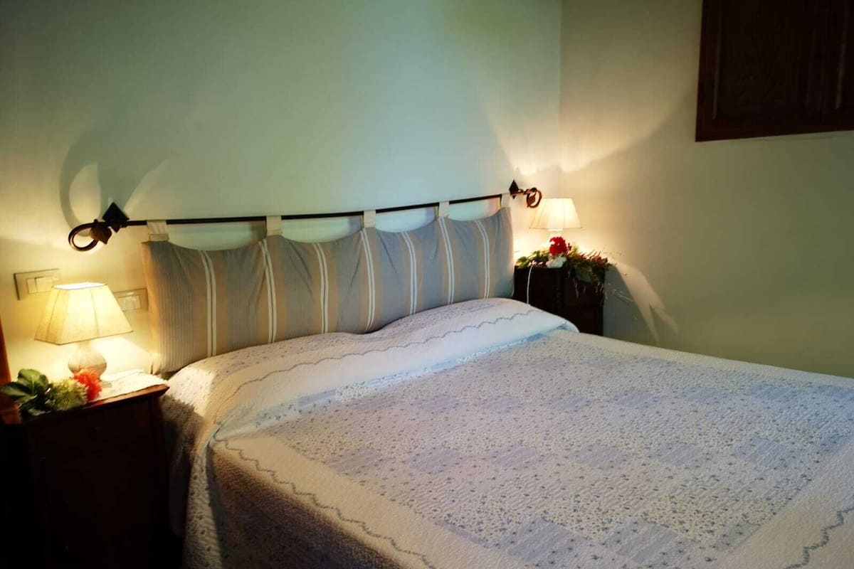 Una cama bien tendida con una manta blanca, una lámpara de noche y una mesita con una flor.