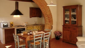 A kitchen with a brick arch, wooden cabinets, a dining table with chairs, and a cabinet with glass doors. - Frateria Di San Benedetto (Montepulciano)