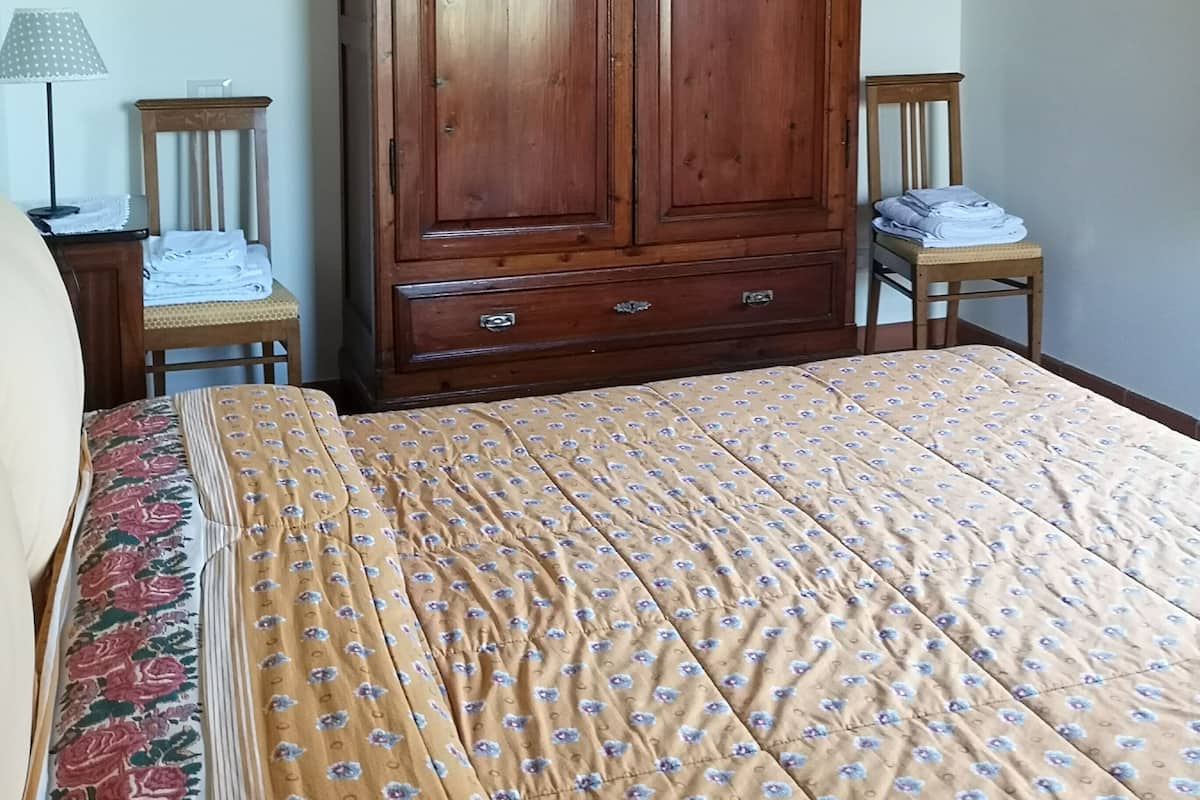 Un dormitorio con un armario grande de madera, una cama con colcha estampada, una silla con toallas dobladas y una lámpara sobre una mesita de noche.
