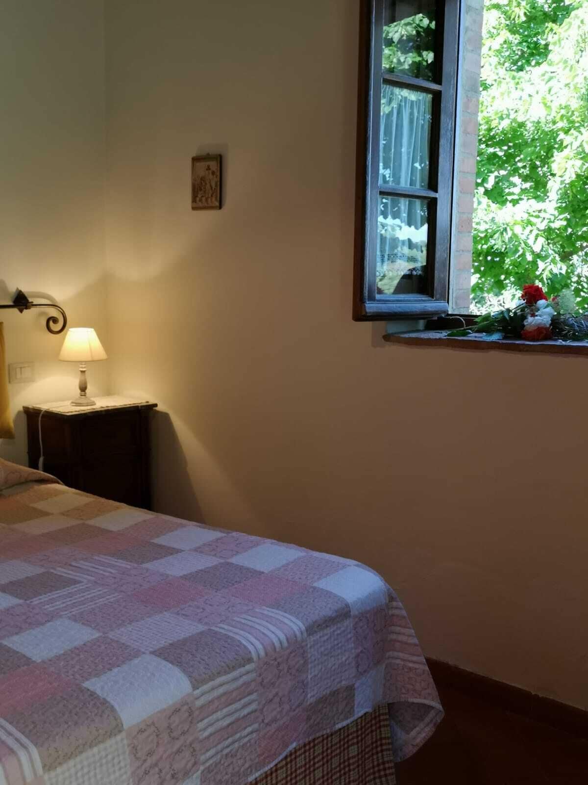Een slaapkamer met een bed, een nachtkastje met een lamp, een raam met uitzicht op groen en een klein schilderij aan de muur.