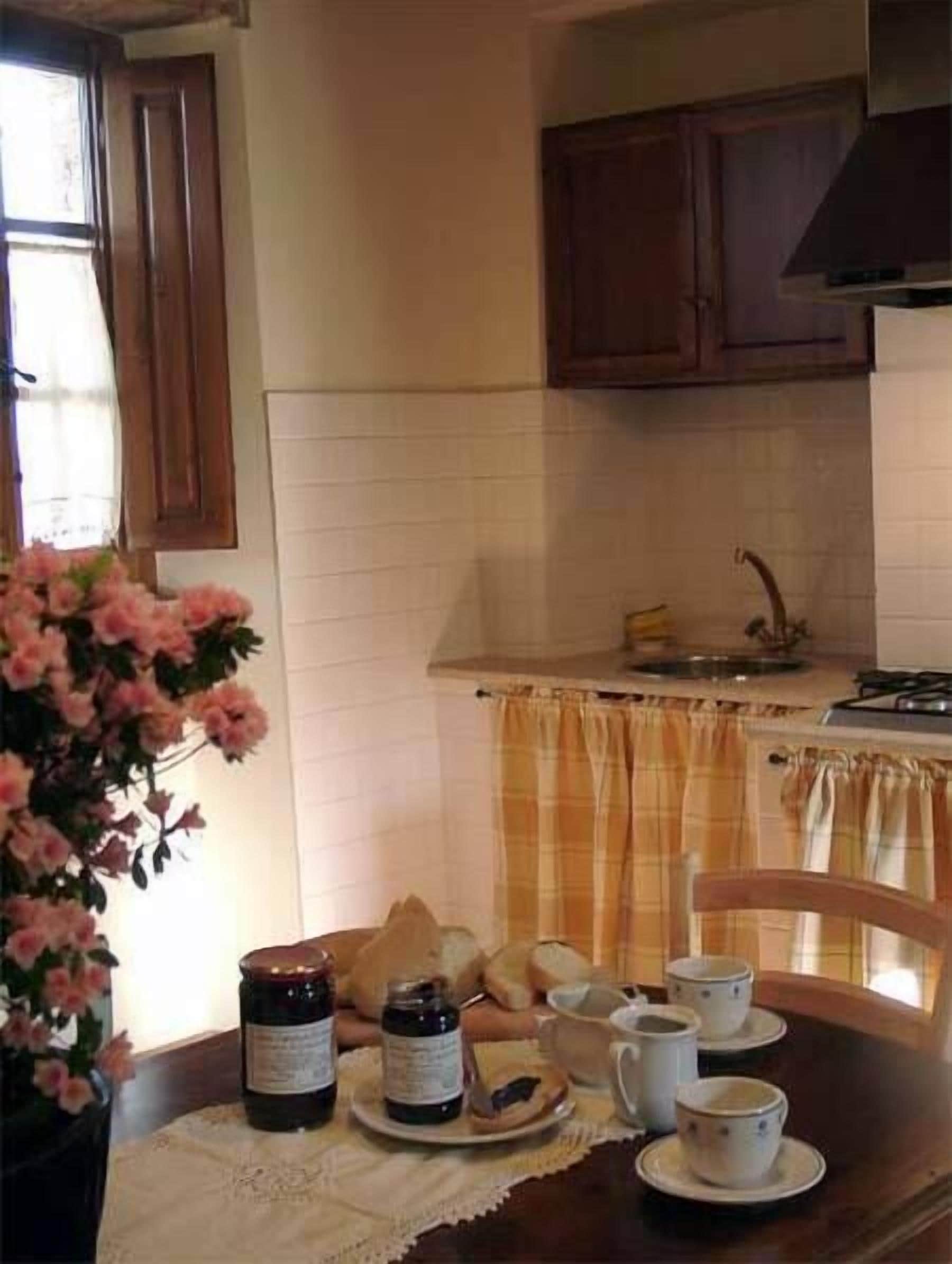 Une cuisine avec des meubles de rangement en bois, une fenêtre avec des rideaux et une table mise avec des tasses, un pot et du pain.