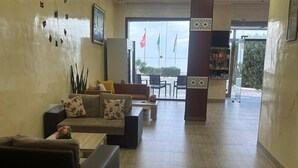 A lobby with a sofa, a coffee table, and a dining area with chairs and a table. There are flags outside the entrance. - Hôtel Al Baraka (Fatou)