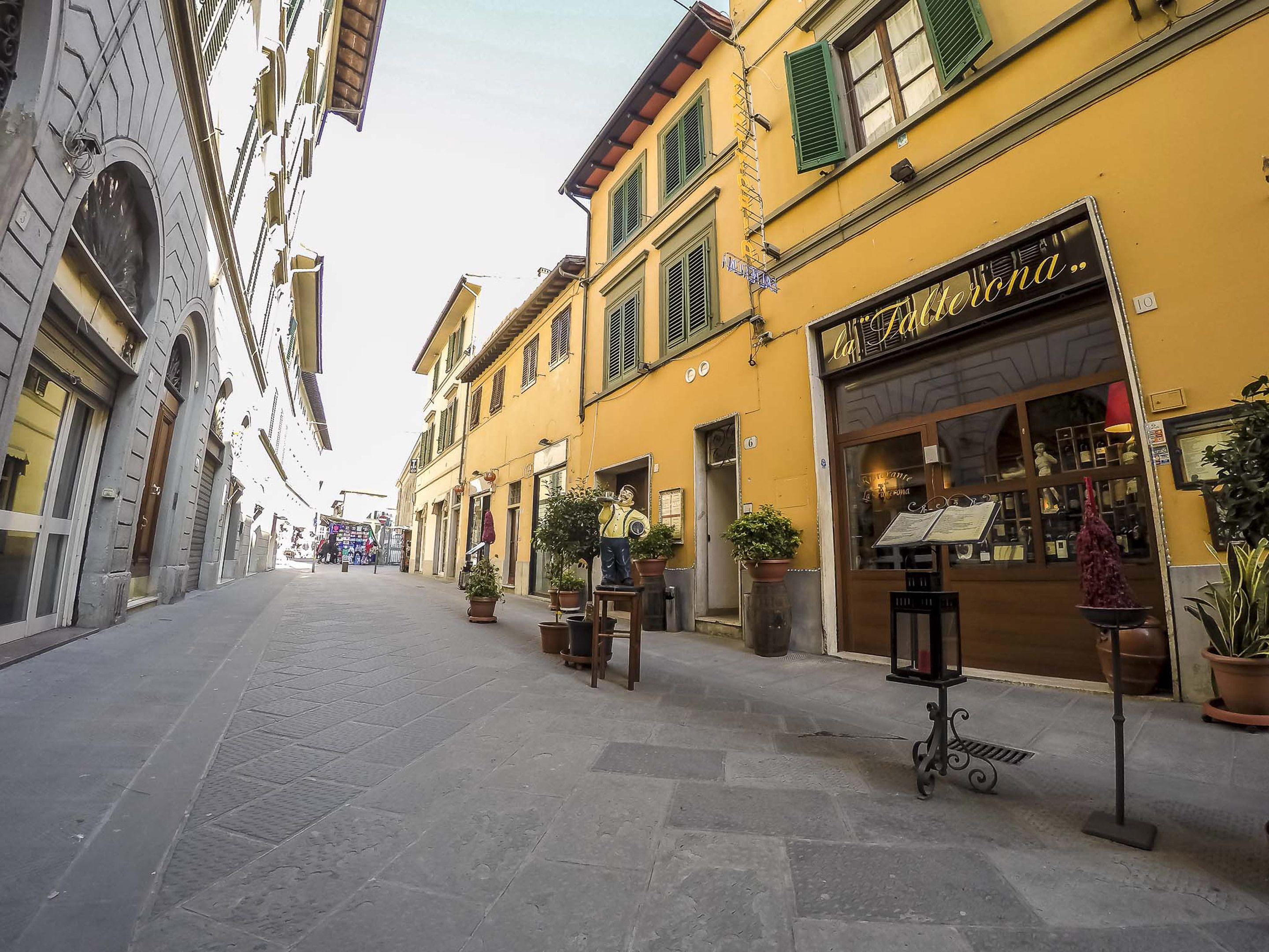 A narrow street with yellow buildings, a restaurant named "La Terza", and potted plants along the sidewalk.