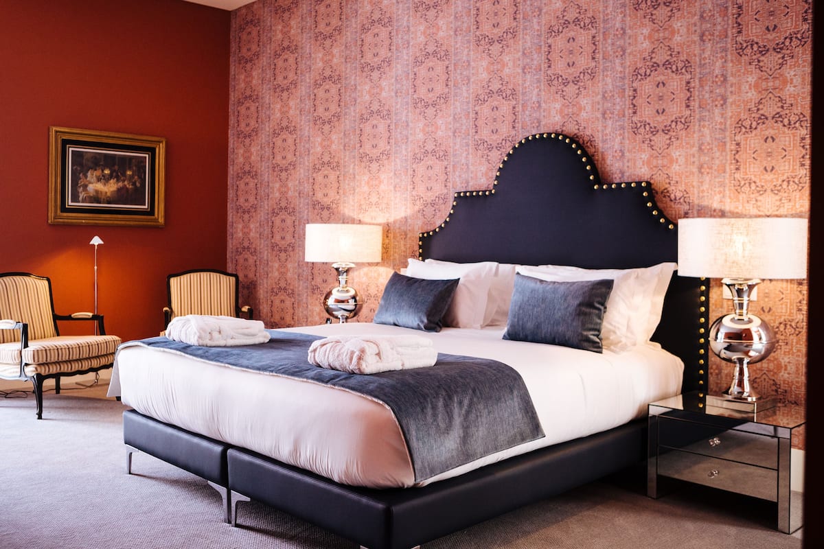 Een opgemaakt bed met een donker hoofdbord, twee bedlampjes en behang met een patroon.