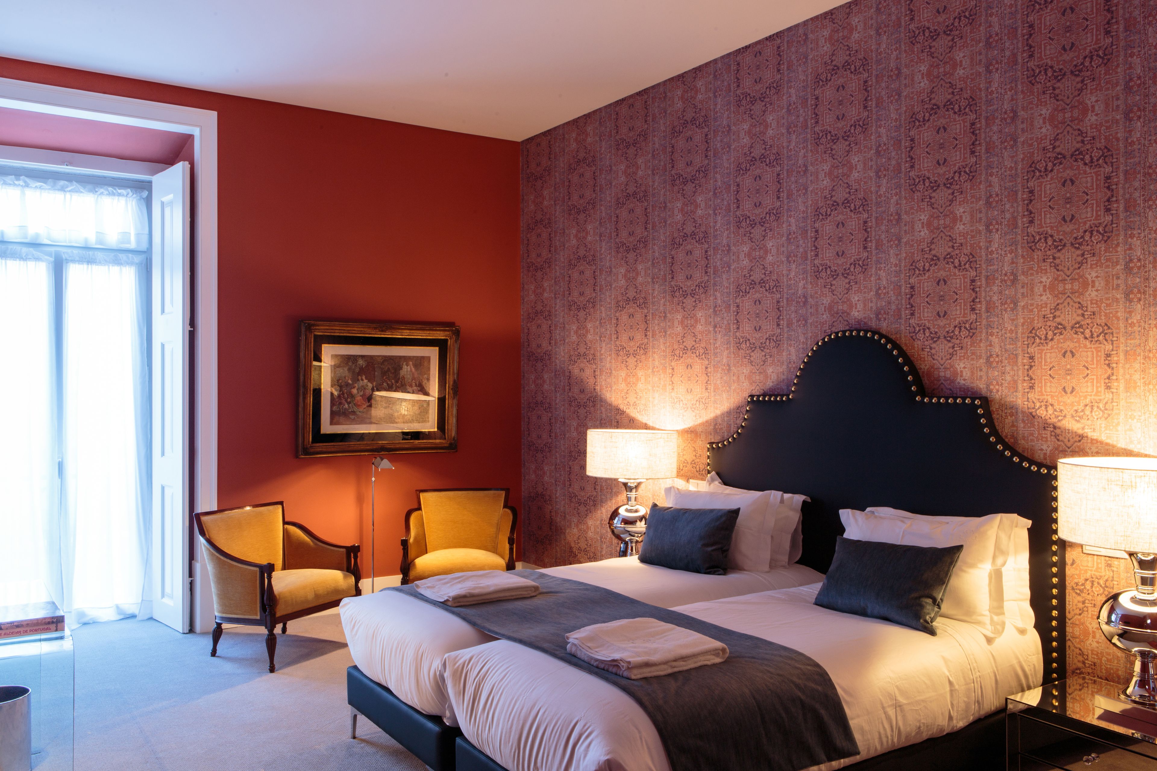Een hotelkamer met een groot bed, twee stoelen, een ingelijst schilderij aan de muur en behang met een patroon.