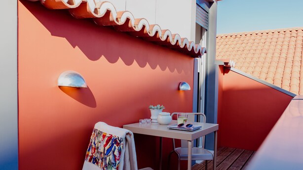 A balcony with a red wall, a white table, a chair, a towel, a potted plant, and a small table lamp.