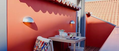 A balcony with a red wall, a white table, a chair, a towel, a potted plant, and a small table lamp.